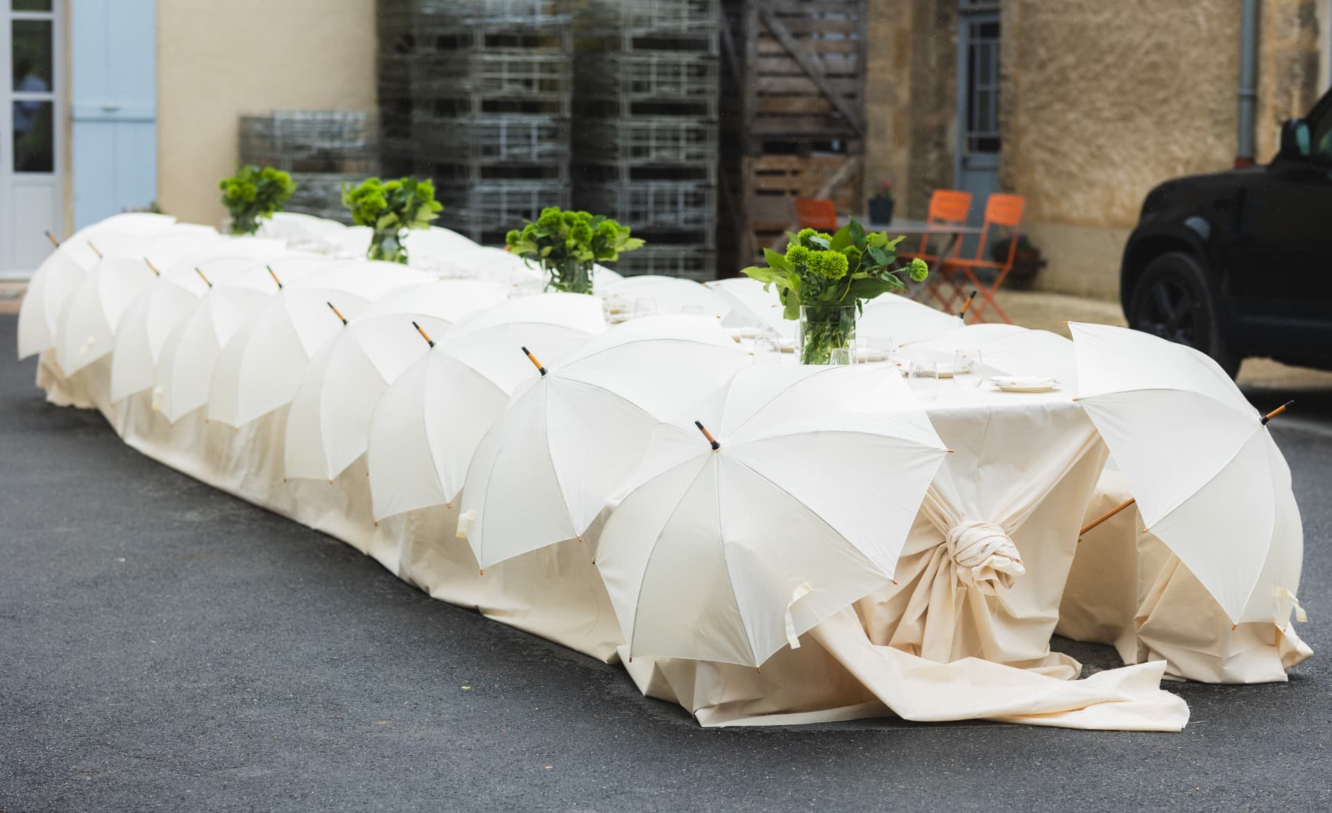 A long HOUSEWINE À TABLE table beneath a row of white parasols in a courtyard.